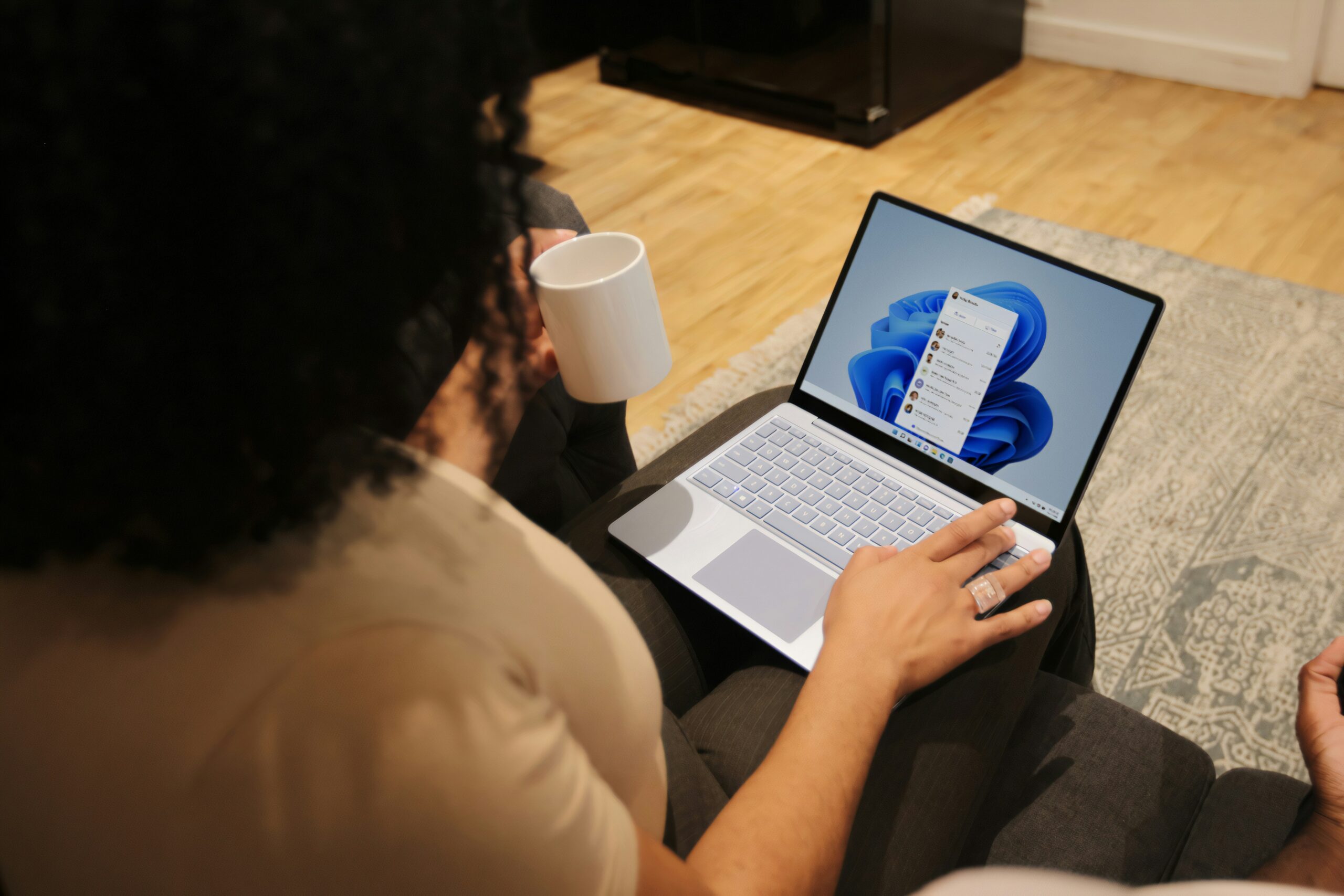 Persona que sostiene una taza blanca y utiliza un ordenador portátil sobre su regazo, en el que se muestra el sistema operativo Windows 11.