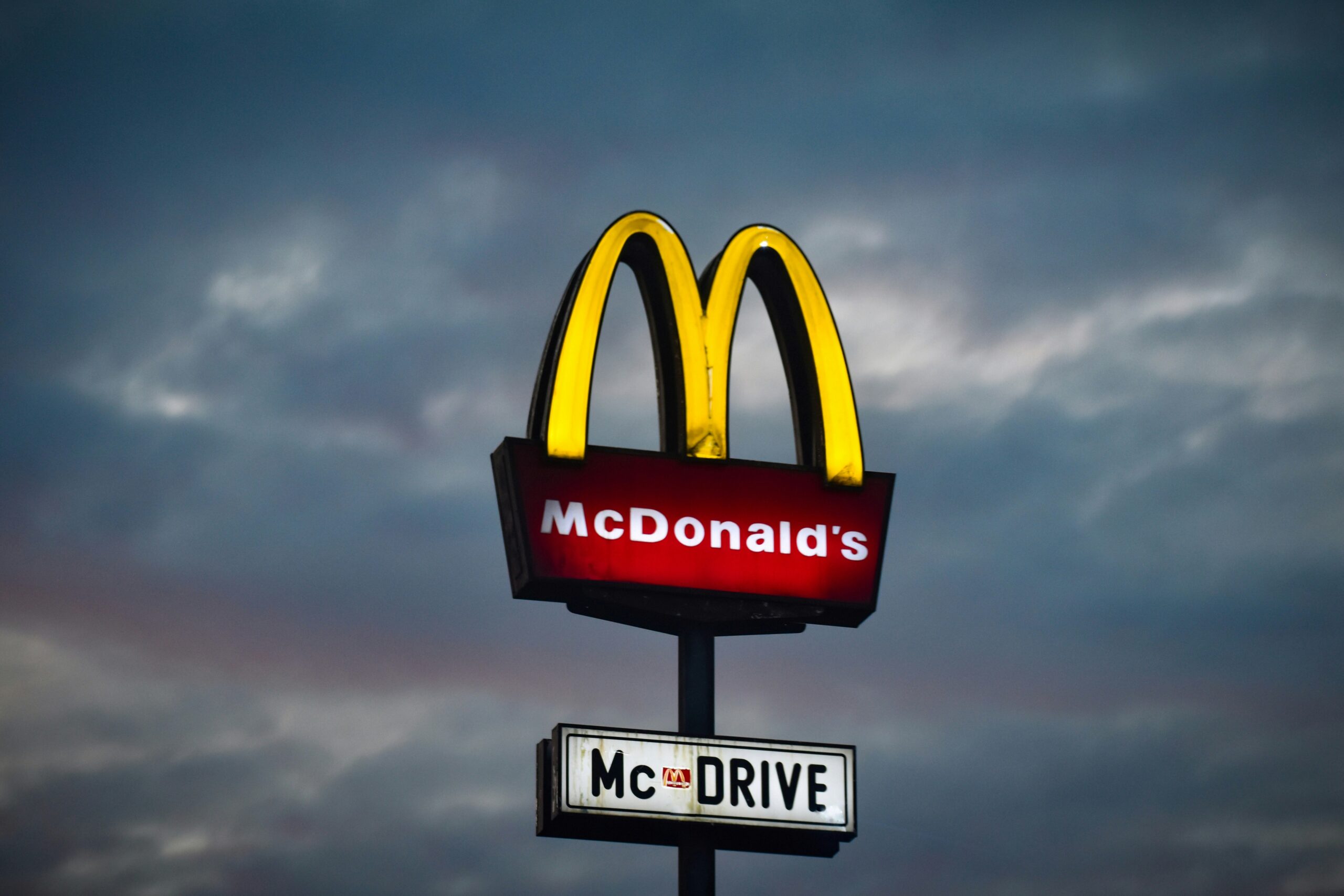 Un letrero de restaurante McDonald's con arcos dorados iluminados y "McDRIVE" visible, sobre un cielo nublado al atardecer.