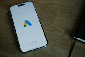 Un smartphone muestra el logotipo de la aplicación Google Ads en su pantalla, apoyado sobre una superficie de madera junto a un cuaderno negro cerrado.
