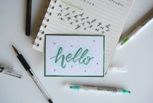 Una nota manuscrita dice "hola" en caligrafía, rodeada de bolígrafos, rotuladores y un cuaderno para practicar el alfabeto cursivo.