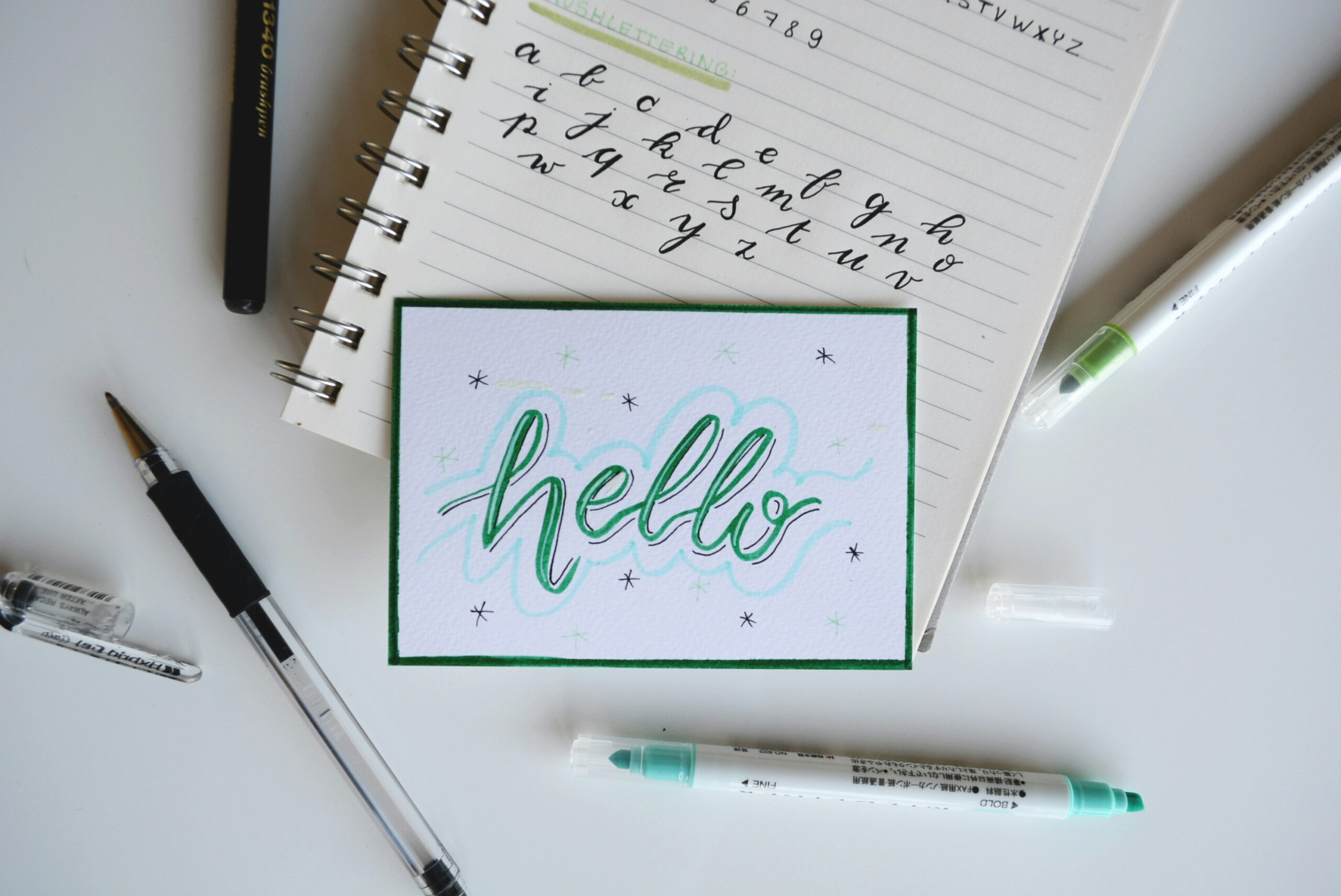 Una nota manuscrita dice "hola" en caligrafía, rodeada de bolígrafos, rotuladores y un cuaderno para practicar el alfabeto cursivo.