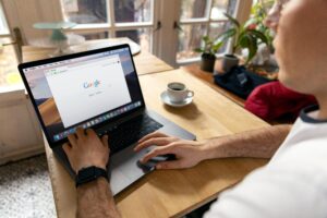 Persona utilizando un ordenador portátil sobre una mesa de madera con la página de inicio de Google abierta; una taza de café, plantas en macetas y una chaqueta también están sobre la mesa cerca de una ventana.