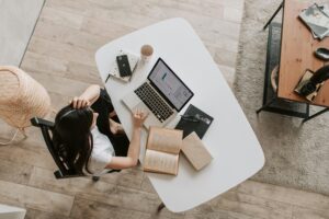 Una persona sentada en un escritorio blanco con un ordenador portátil, un libro abierto, cuadernos, un smartphone y una taza, vista desde arriba en un entorno doméstico.