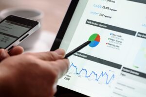Una persona apunta a una tableta que muestra tablas, gráficos y datos analíticos de un sitio web, mientras sostiene un smartphone en la otra mano.