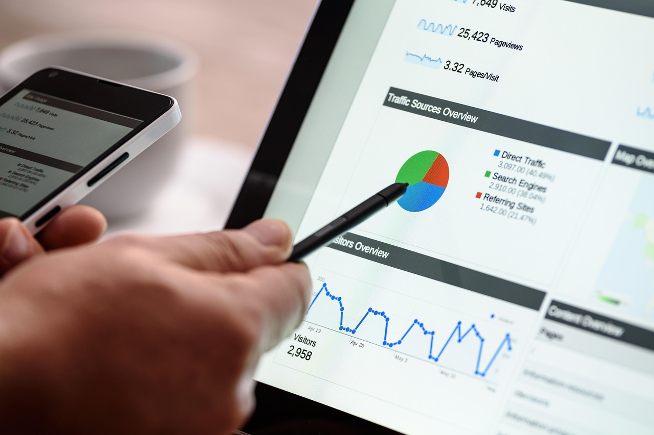 Una persona apunta a una tableta que muestra tablas, gráficos y datos analíticos de un sitio web, mientras sostiene un smartphone en la otra mano.