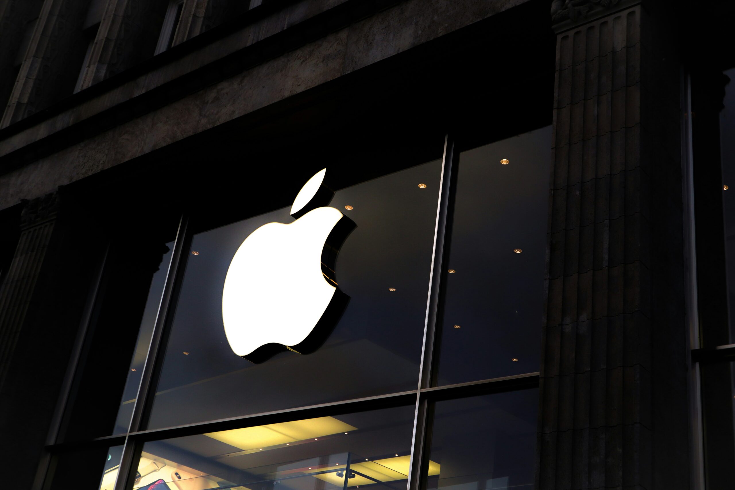 Gran logotipo de Apple iluminado en el escaparate de cristal de una Apple Store, con parte del exterior del edificio visible.