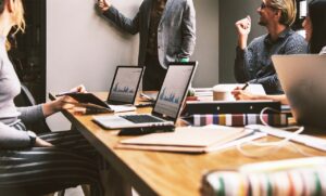 En una sala de reuniones, los asistentes se sientan alrededor de una mesa con ordenadores portátiles y cuadernos, mientras una persona presenta de pie los datos que aparecen en la pantalla de un portátil.