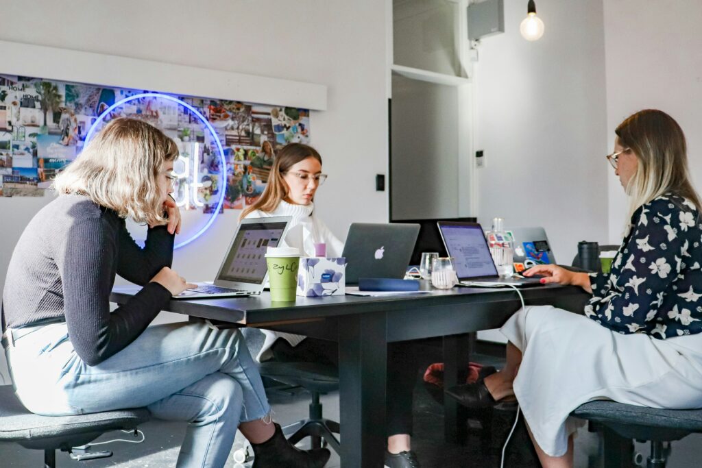Tres mujeres se sientan ante una mesa negra en una oficina bien iluminada, trabajando en ordenadores portátiles con cuadernos, bebidas y otros objetos sobre la mesa.