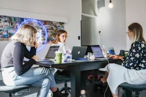 Tres mujeres se sientan ante una mesa negra en una oficina bien iluminada, trabajando en ordenadores portátiles con cuadernos, bebidas y otros objetos sobre la mesa.
