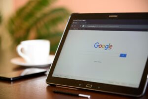 Una tableta Samsung que muestra la página de inicio de Google sobre una mesa de madera, con un lápiz óptico, un smartphone y una taza blanca de fondo.