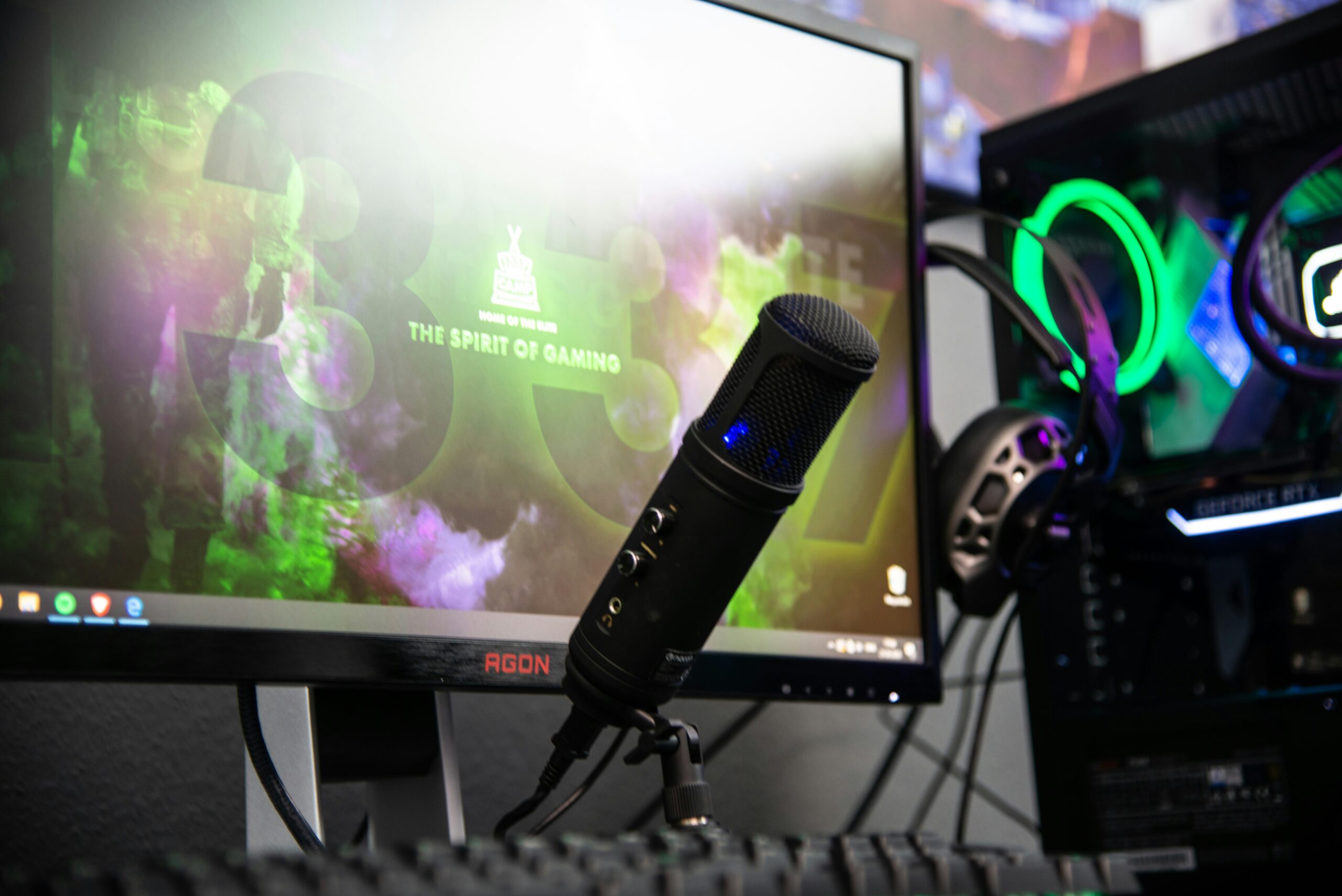 Una configuración de escritorio con un monitor que muestra un fondo de colores, un micrófono negro en un soporte, componentes de PC iluminados con RGB y un teclado en primer plano.