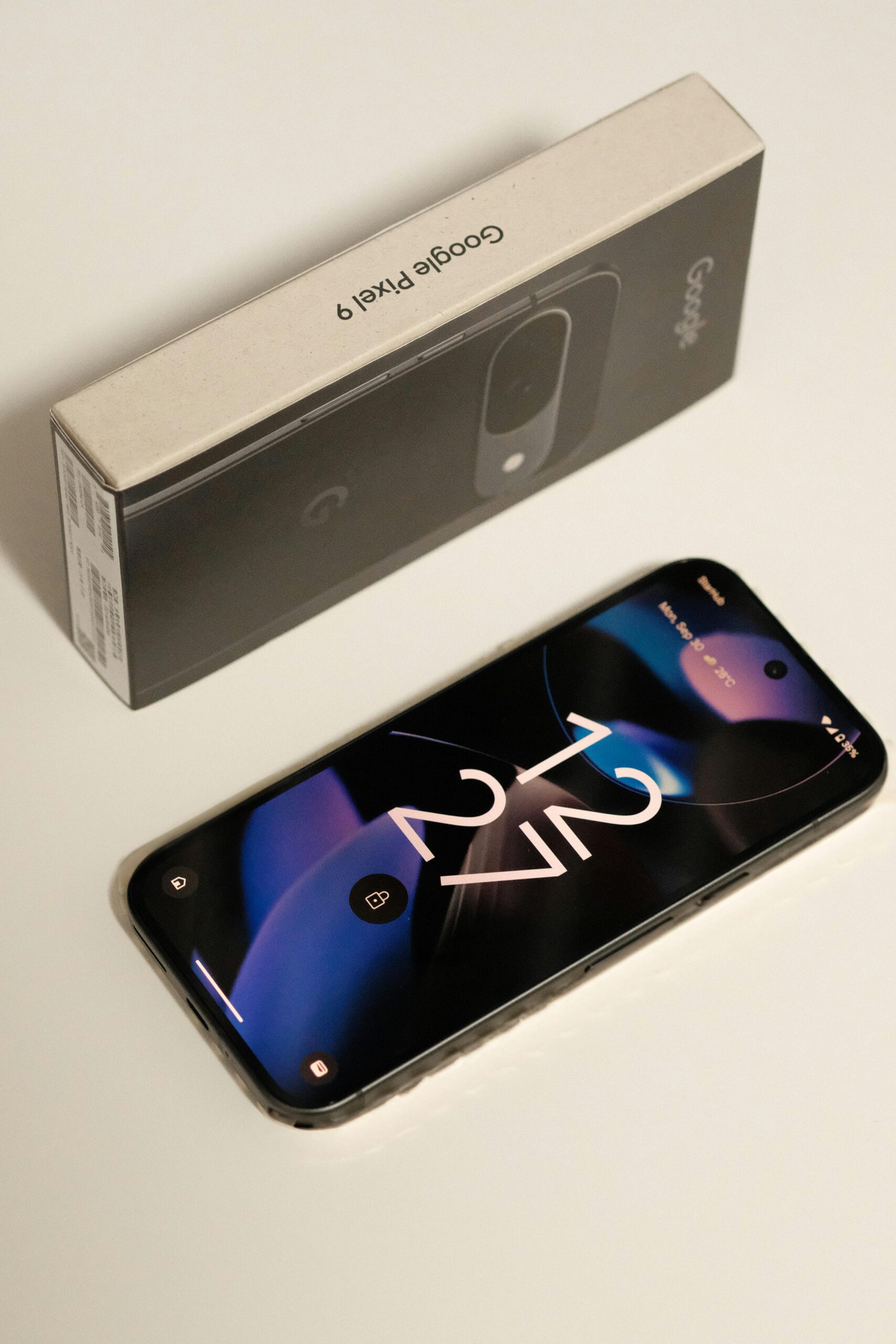 Un smartphone Google Pixel 9 yace boca arriba sobre una superficie blanca junto a su caja de embalaje original.