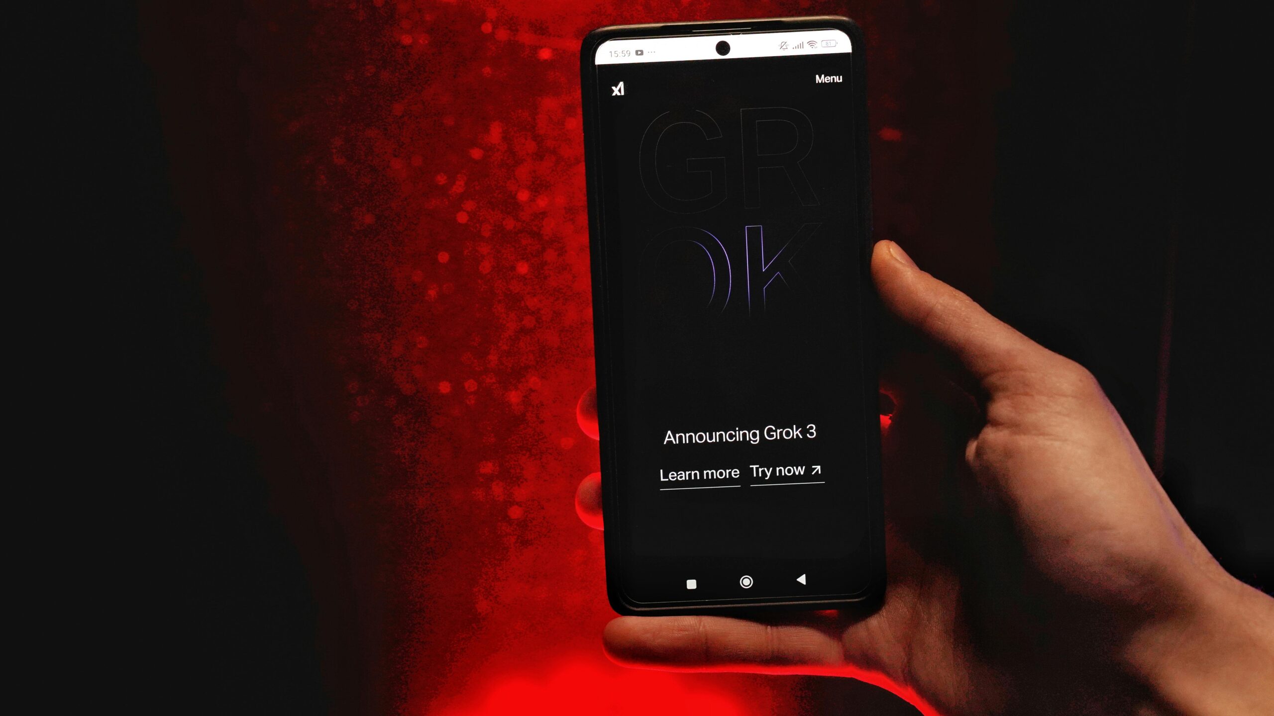 Una mano sujeta un smartphone que muestra una pantalla oscura con el texto "Announcing Grok 3" y las opciones "Learn more" y "Try now", sobre un fondo iluminado en rojo.
