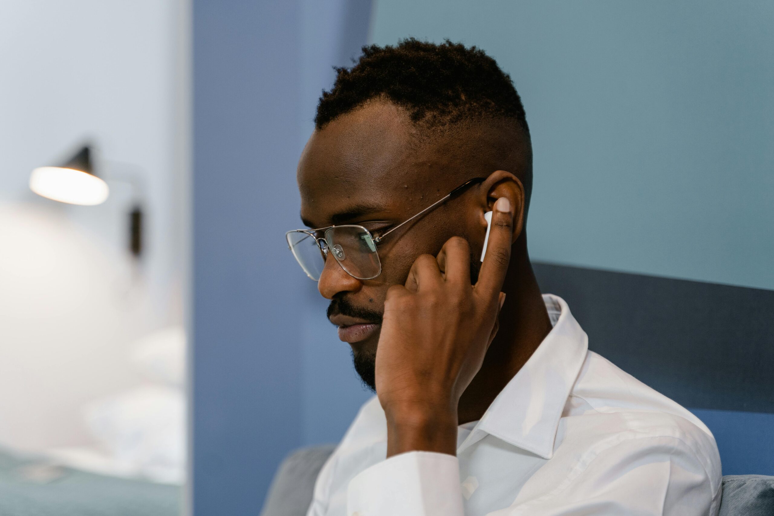 Un hombre con gafas y camisa blanca escucha algo en unos auriculares inalámbricos tocándose la oreja mientras está sentado en un interior.