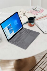 Un portátil gris que muestra el menú de inicio de Windows 10 se sienta en una mesa blanca redonda con cuadernos abiertos, papeles sueltos, bolígrafos y una taza.