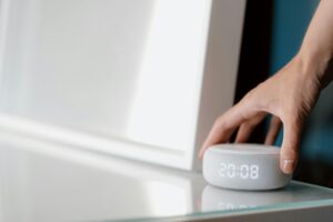 Una mano alcanza un altavoz inteligente blanco con un reloj digital que muestra las 20:08, colocado sobre una superficie de cristal.