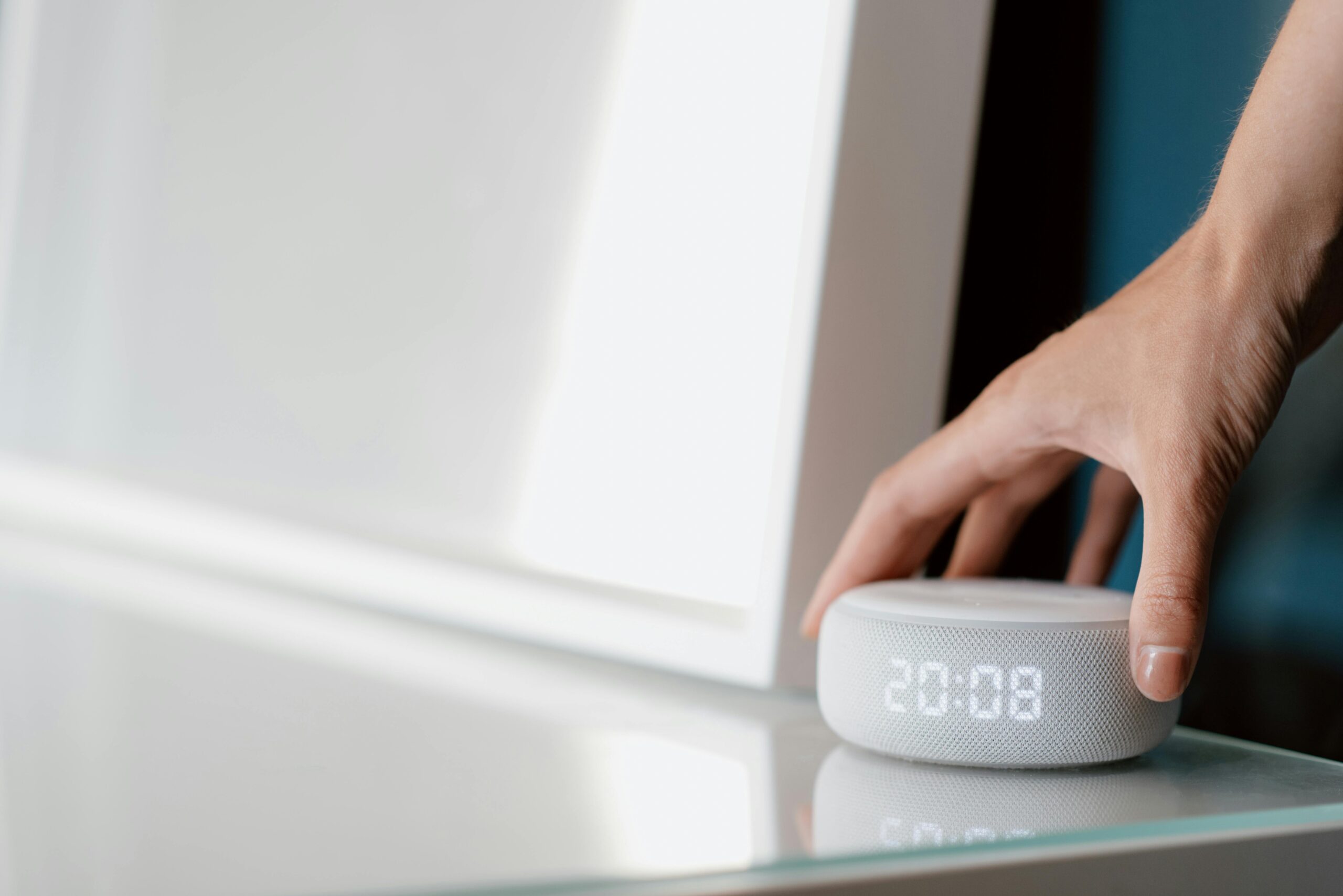 Una mano alcanza un altavoz inteligente blanco con un reloj digital que muestra las 20:08, colocado sobre una superficie de cristal.