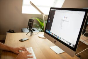 Persona escribiendo en un teclado en un escritorio de madera con un iMac que muestra la búsqueda de Google, un micrófono, una lámpara, un teléfono y una planta también están en el escritorio.