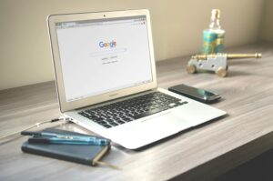 Un ordenador portátil con la página de inicio de Google abierta descansa sobre un escritorio junto a un smartphone, un cuaderno con bolígrafos, auriculares, una botella y un pequeño cañón decorativo.