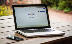 Un MacBook Pro sobre una mesa de madera muestra la página de inicio de Google; un smartphone descansa junto al portátil.