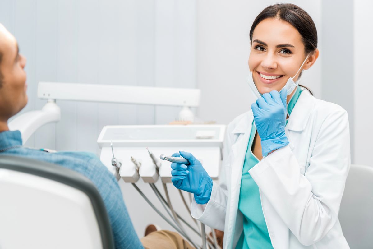 Un dentista con guantes y bata blanca sonríe sentado junto a un paciente en una clínica dental local, con un instrumento dental en la mano, haciendo hincapié en la atención personalizada y la importancia de las estrategias seo locales para la visibilidad en la comunidad.