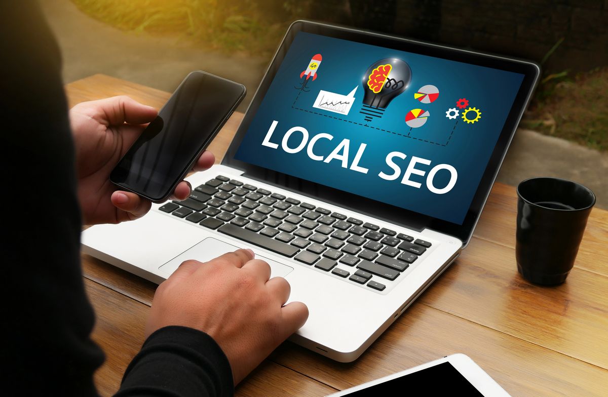 Una persona que utiliza un teléfono inteligente y un ordenador portátil en cuya pantalla aparecen las palabras "SEO local", rodeada de gráficos y diagramas, está sentada a una mesa de madera con una taza de café y una tableta.