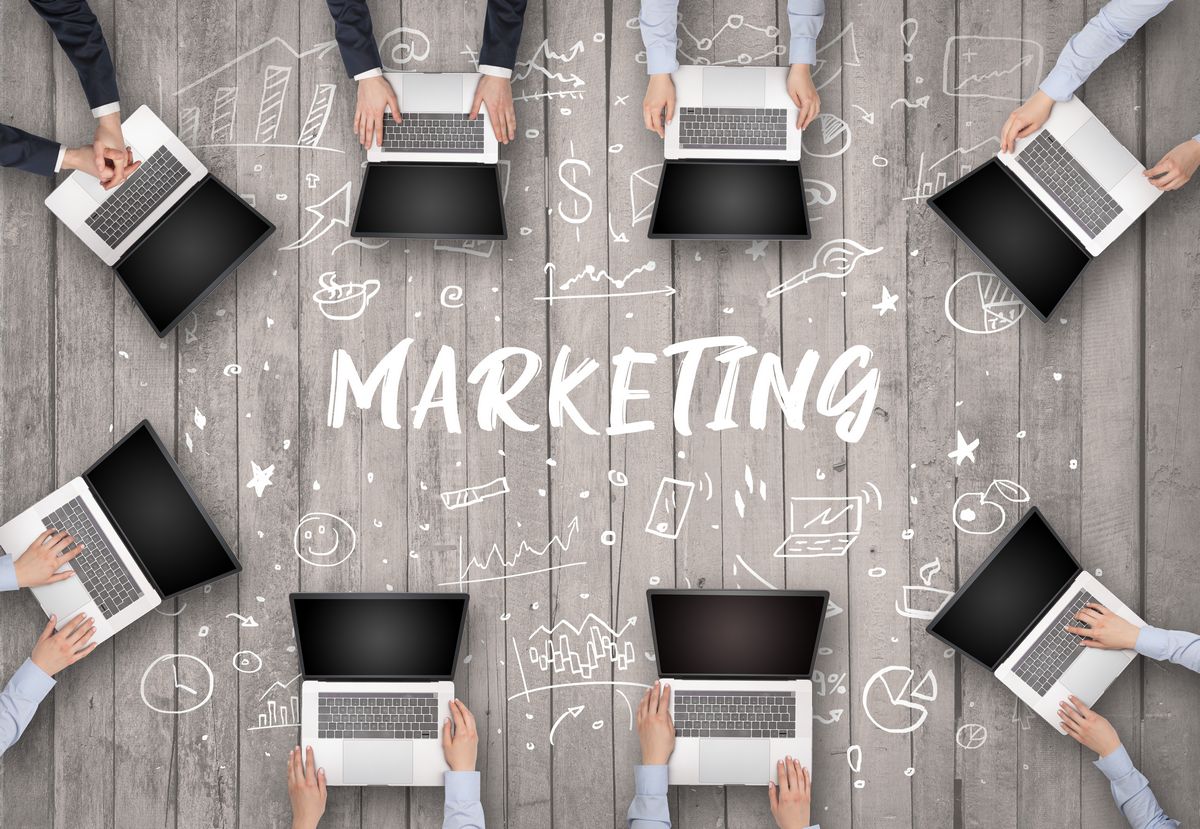 Ocho personas trabajando con ordenadores portátiles dispuestas en círculo sobre un suelo de madera, con la palabra "MARKETING", seo en Tijuana, y bocetos relacionados con negocios dibujados en el centro.