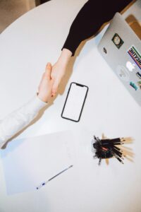 Dos personas se dan la mano sobre una mesa blanca en la que se ven un ordenador portátil, un teléfono, papel, bolígrafo y un portalápices.
