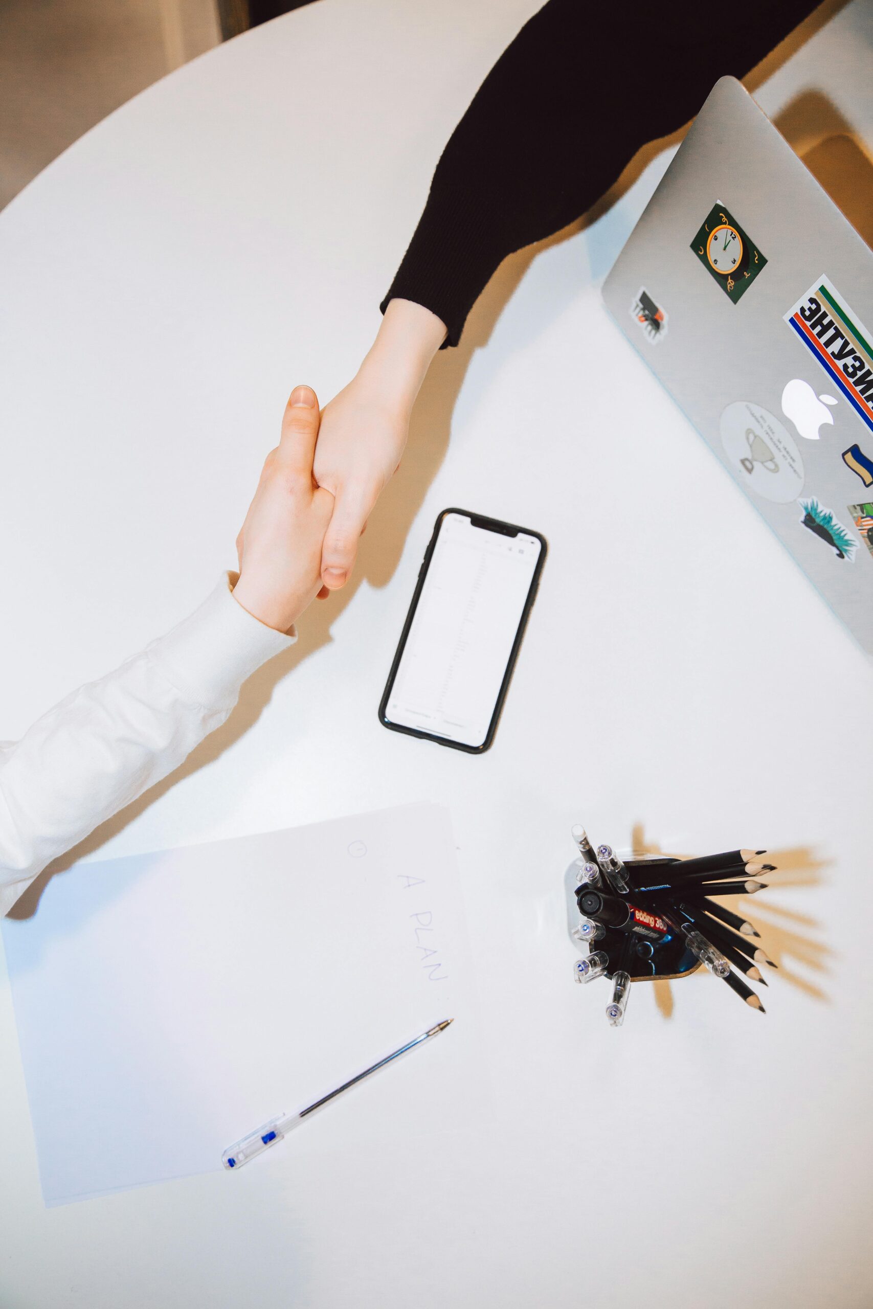 Dos personas se dan la mano sobre una mesa blanca en la que se ven un ordenador portátil, un teléfono, papel, bolígrafo y un portalápices.