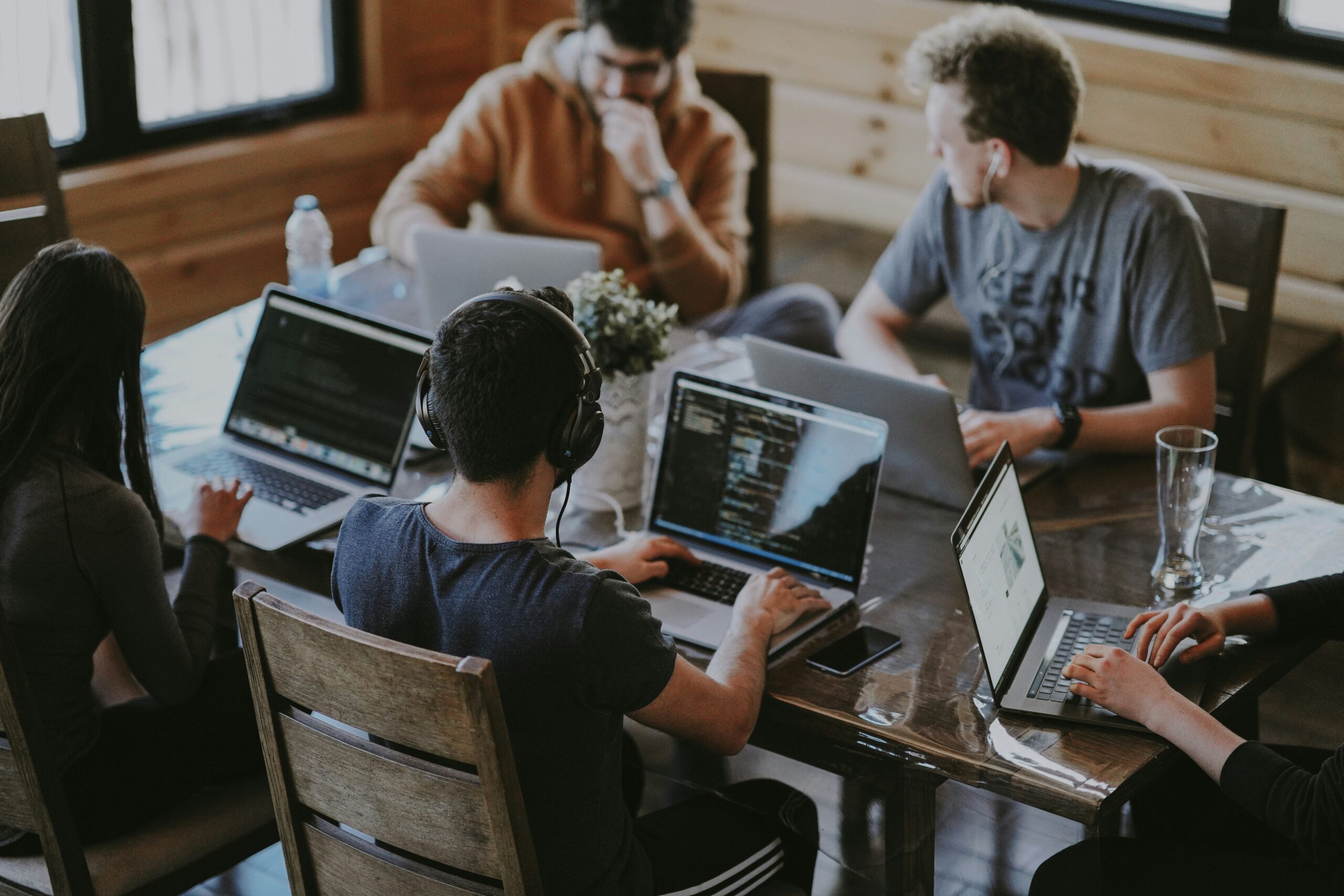 Cinco personas se sientan alrededor de una mesa a trabajar con sus portátiles, algunas con auriculares, en un ambiente informal de interior con luz natural.