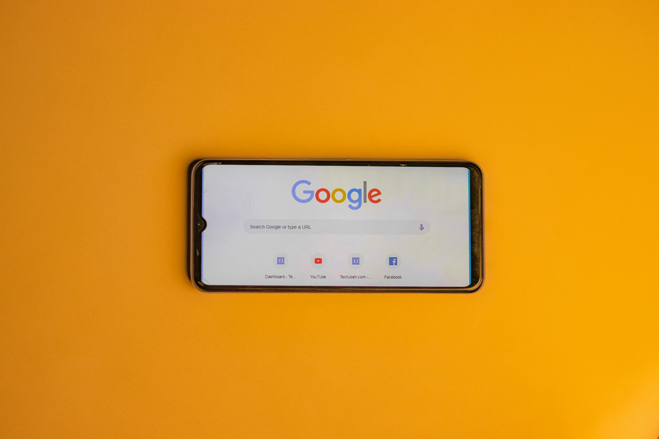 Sobre una superficie lisa de color naranja se encuentra un smartphone que muestra en su pantalla la página de inicio de la búsqueda de Google.