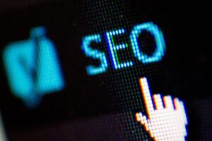 En la pantalla de un ordenador aparece la palabra "SEO" junto al icono de un cursor en forma de mano, lo que sugiere que se centra en la optimización de los motores de búsqueda.