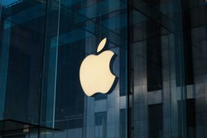 Gran logotipo de Apple iluminado en el exterior de cristal de una tienda Apple, con reflejos de edificios al fondo.