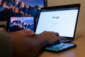 Persona que utiliza un ordenador portátil con la página de búsqueda de Google abierta; sobre el escritorio hay también un smartphone y una tableta con un fondo de pantalla de paisaje montañoso.