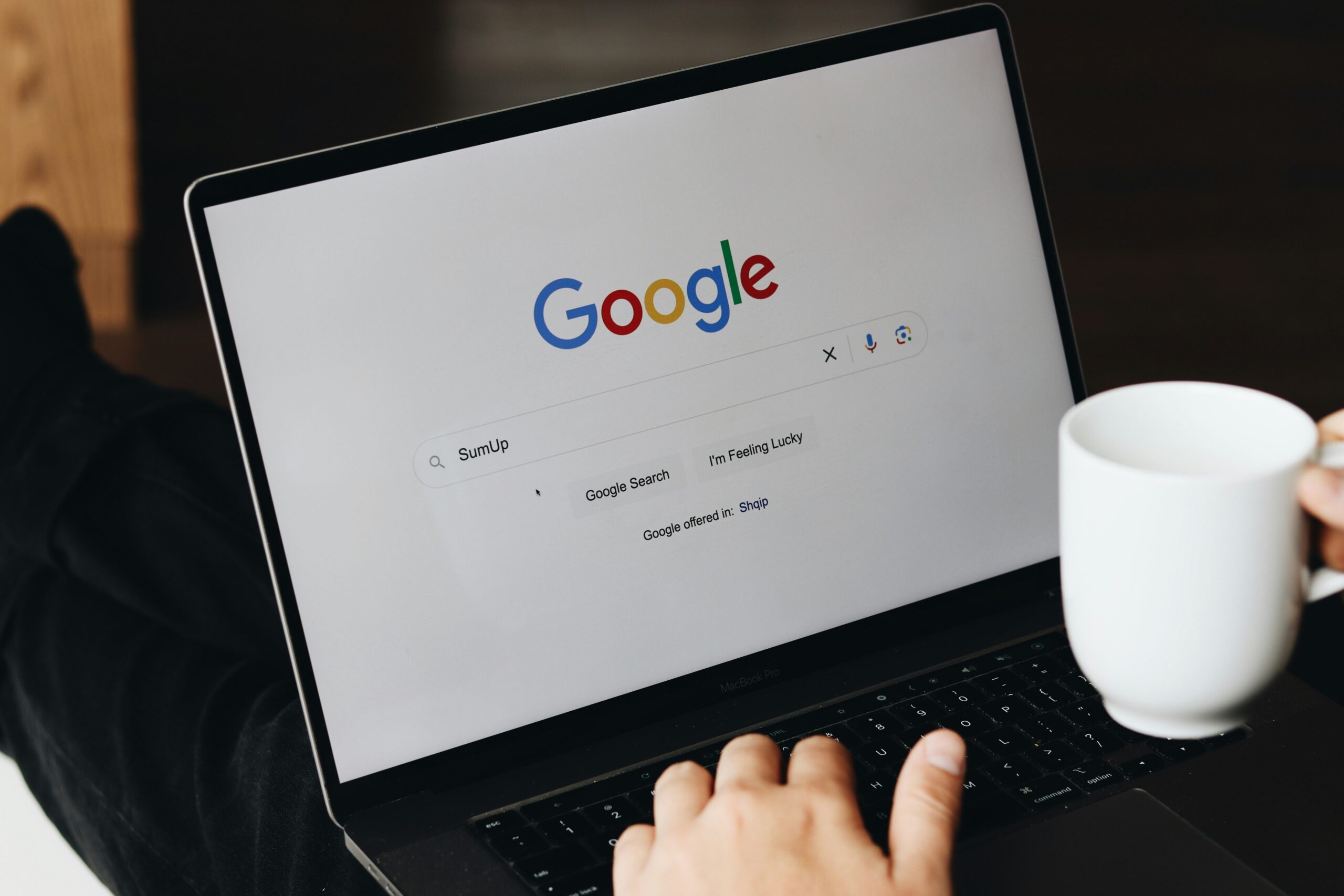 Una persona con una taza blanca utiliza un ordenador portátil que muestra la página principal de Google, con el término de búsqueda "SumUp" escrito en la barra de búsqueda.