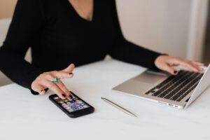 Persona utilizando un smartphone mientras está sentada en un escritorio con un portátil abierto y un bolígrafo sobre una superficie blanca.
