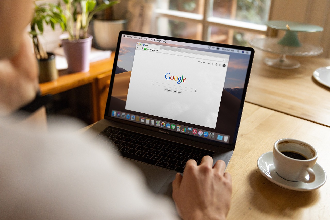 Una persona utiliza un ordenador portátil que muestra la página de inicio de Google en una mesa de madera con una taza de café, plantas en macetas y una ventana al fondo.