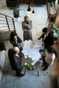 Cinco personas vestidas con traje de negocios se sientan alrededor de una mesa con papeles, planos de construcción y un ordenador portátil, enfrascadas en una discusión en una oficina moderna.