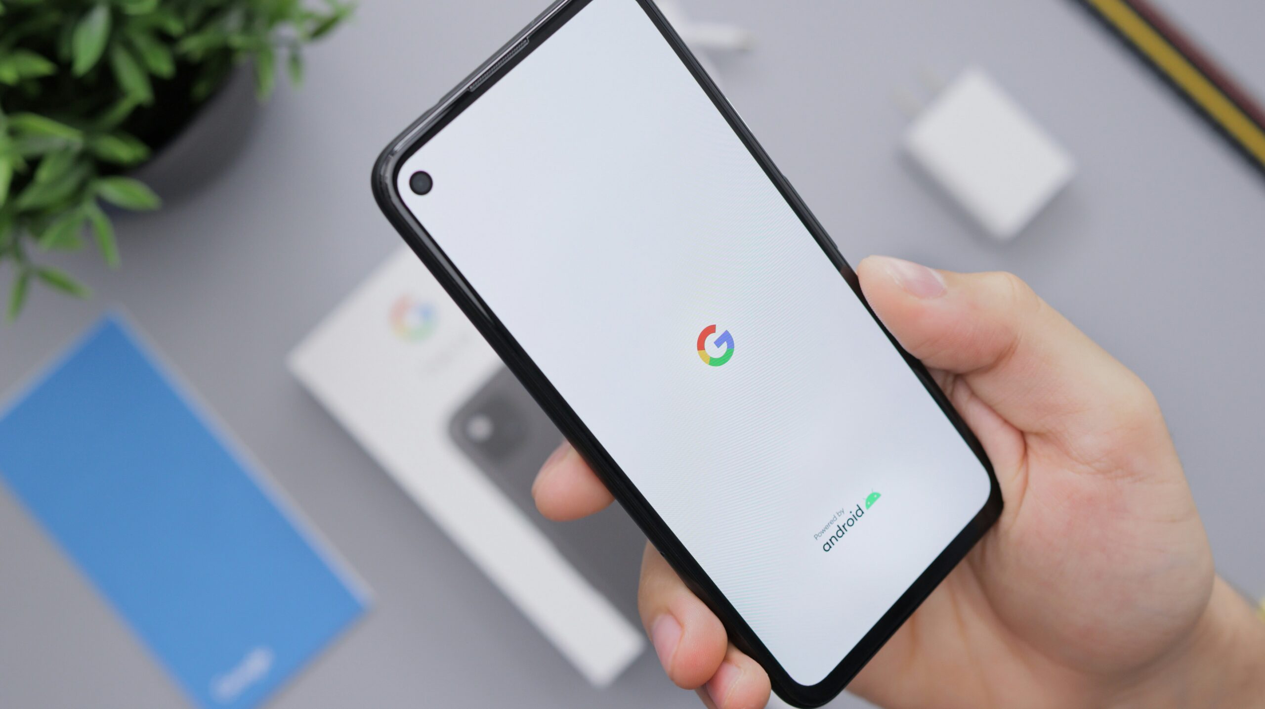 Una mano sostiene un smartphone que muestra los logotipos de Google y Android en su pantalla de inicio; en el fondo se ven un cargador, una caja y un folleto.