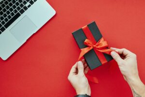 Persona que sostiene una caja de regalo negra con un lazo rojo junto a un ordenador portátil sobre una superficie roja.