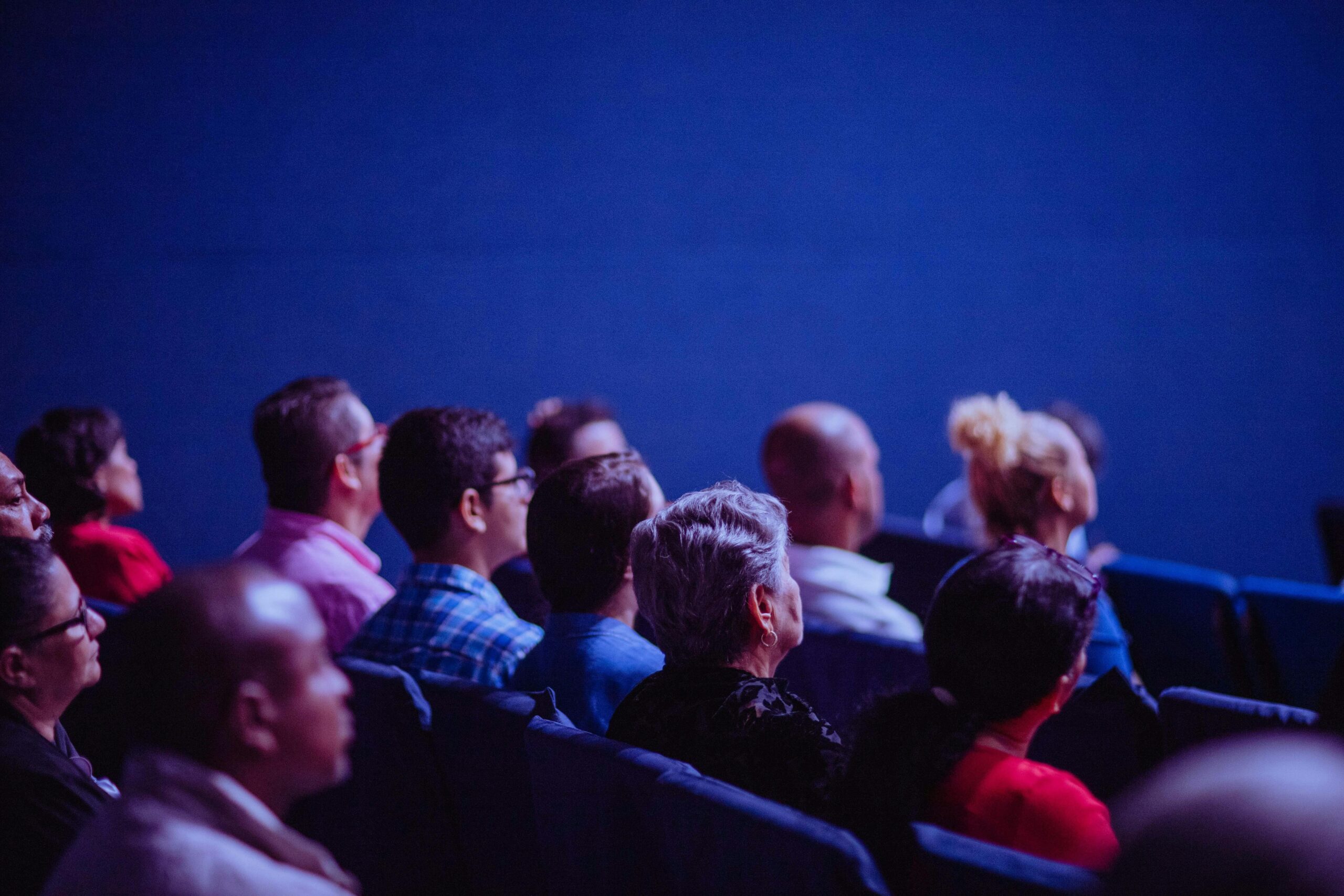 Miembros del público sentados en filas, mirando hacia delante y observando atentamente una presentación o actuación que no se ve en una sala poco iluminada.