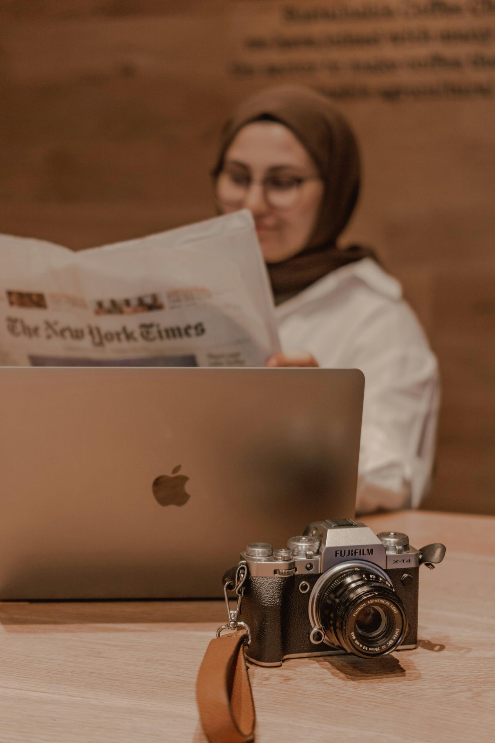 Una persona con hiyab está sentada en un escritorio con un ordenador portátil abierto, leyendo el periódico The New York Times; una cámara Fujifilm descansa sobre la mesa en primer plano.