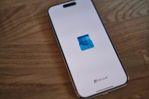 Un smartphone muestra la pantalla de carga de la aplicación Microsoft Outlook, con el logotipo de Outlook sobre fondo blanco. El teléfono descansa sobre una superficie de madera.
