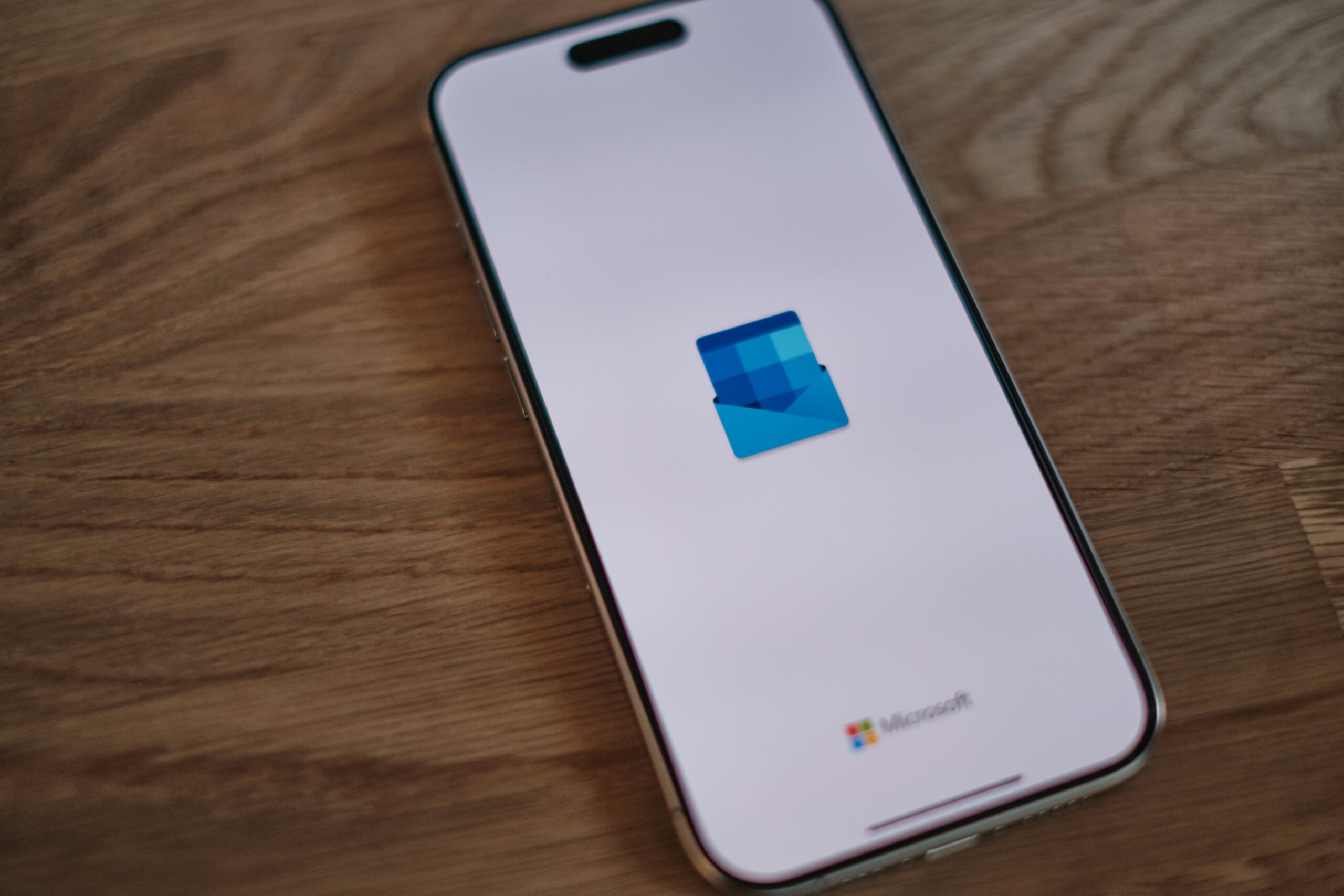 Un smartphone muestra la pantalla de carga de la aplicación Microsoft Outlook, con el logotipo de Outlook sobre fondo blanco. El teléfono descansa sobre una superficie de madera.