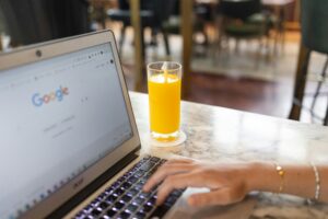 Una persona teclea en un portátil que muestra la página de inicio de Google, con un vaso de zumo de naranja sobre la mesa.