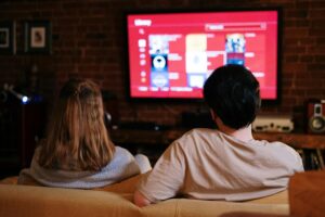 Dos personas se sientan en un sofá frente a una gran pantalla de televisión que muestra el menú de un servicio de streaming en un salón poco iluminado.