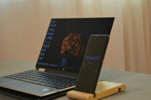 Un ordenador portátil con un fondo de pantalla de flores y un smartphone sobre un soporte de madera están colocados sobre una mesa. En la pantalla del teléfono aparece el texto "MAKE A NON HAPPY.