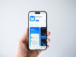 Una mano sostiene un smartphone que muestra la página de la aplicación Bluesky Social en la App Store, con el icono de la aplicación, la descripción y capturas de pantalla.