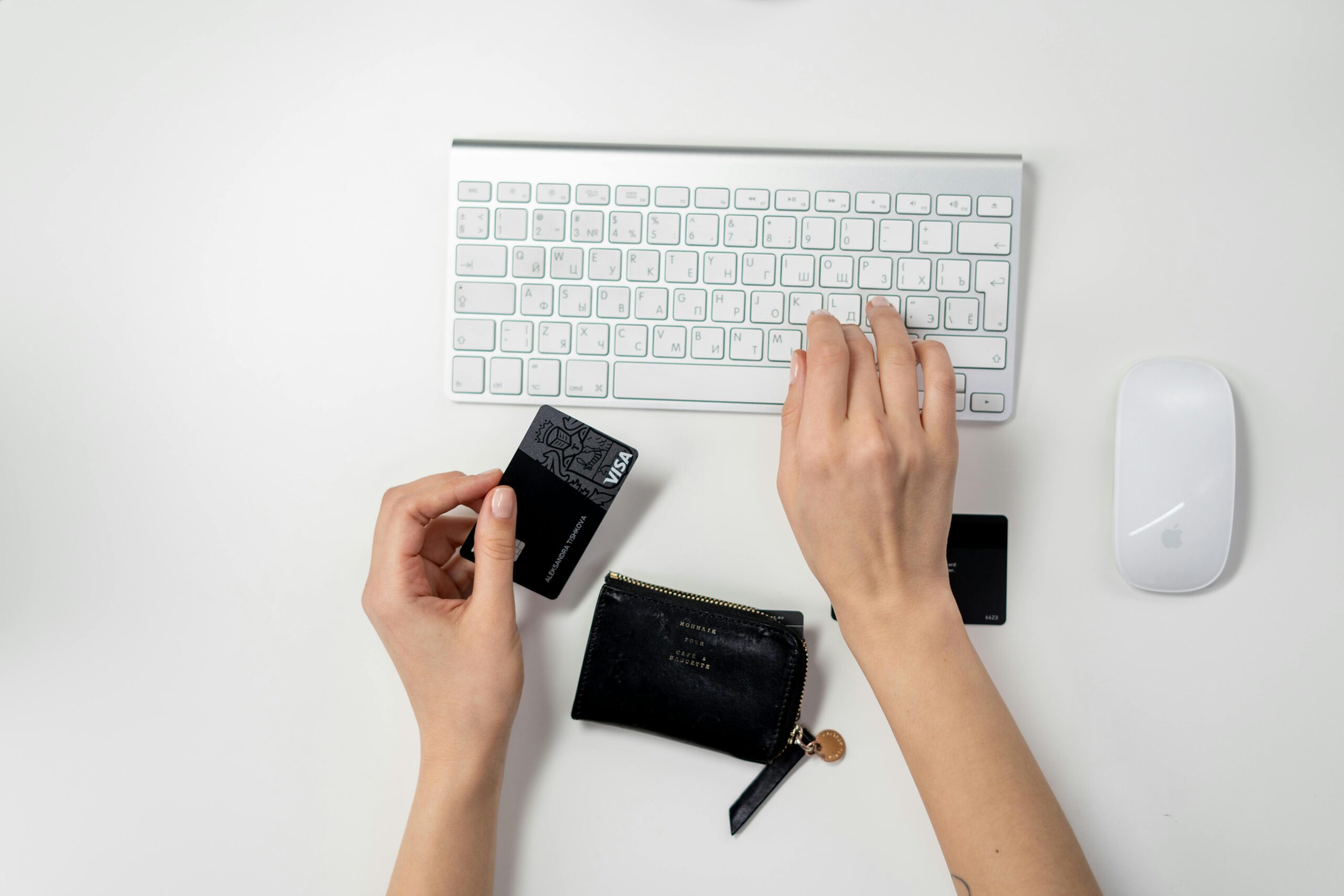 Una persona escribe en un teclado de ordenador blanco con una mano mientras sujeta una tarjeta de crédito negra y una pequeña cartera negra con la otra. Cerca hay un ratón blanco.