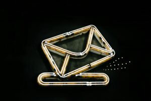 Un letrero de neón con forma de sobre apoyado sobre un clip, ambos perfilados con luz blanca sobre fondo negro.